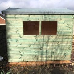 A newly built, light green wooden shed with a pitched roof and two rectangular windows is situated in a garden. The shed sits on bricks, surrounded by a stone patio, potted plants, and a concrete wall on the left.
