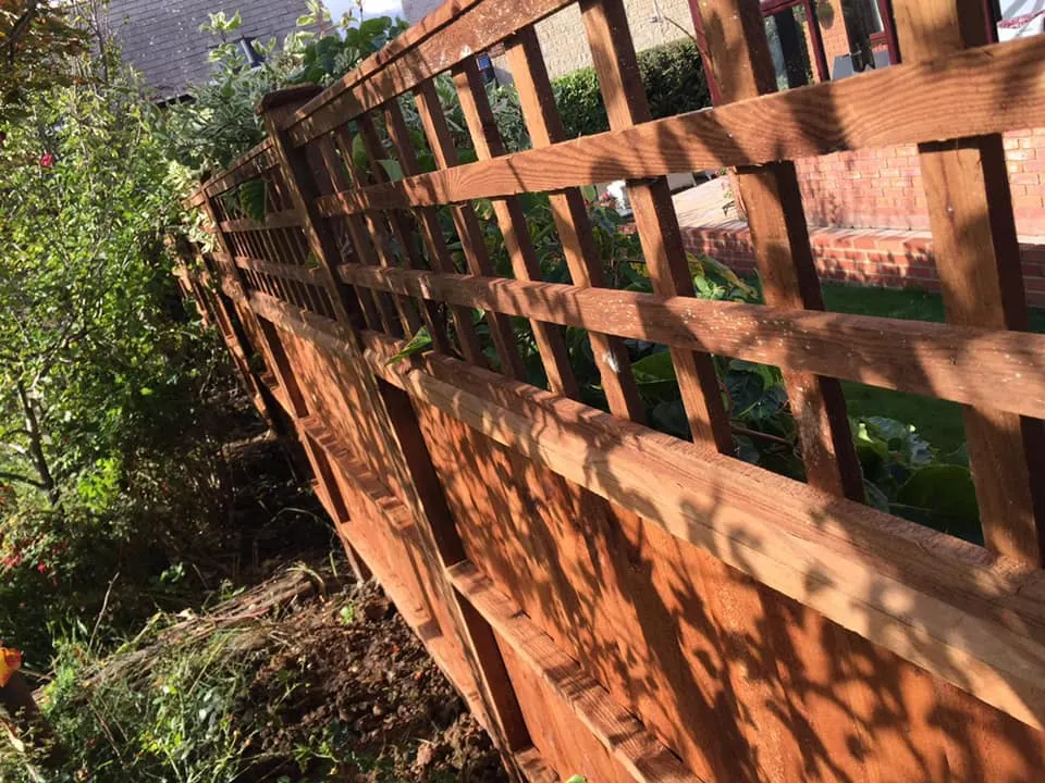 A wooden lattice fence in a garden casts shadows on the ground. Green foliage and trees are visible on the left side, while a red brick building is seen in the background. The scene is sunlit, highlighting the textures of the wood and bricks.
