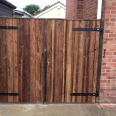 A wooden gate with metal hinges and a latch, set between a brick wall and a wooden fence, leading to a paved pathway. The gate is made of vertical wooden planks with a natural finish. A portion of a house exterior is visible.