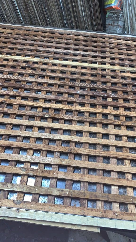 A wooden lattice structure with a grid pattern is displayed. Behind it, there are some bamboo sticks and part of a can with colorful graphics. The lattice consists of thin, interwoven wooden strips.