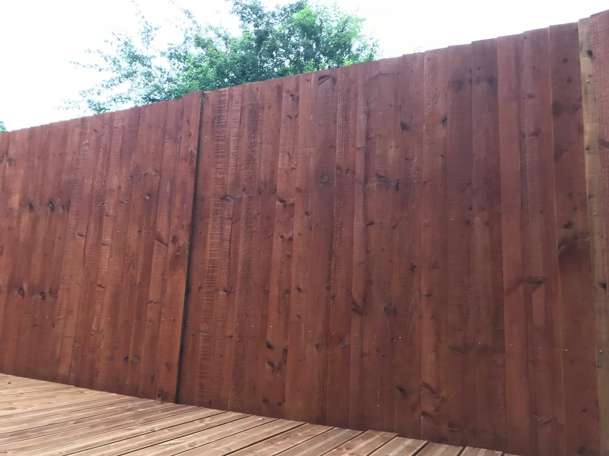 A tall wooden fence made of vertical planks stands on a wooden deck. The fence is a rich brown color and is surrounded by some green foliage in the background. The deck appears to be newly constructed and well-maintained.