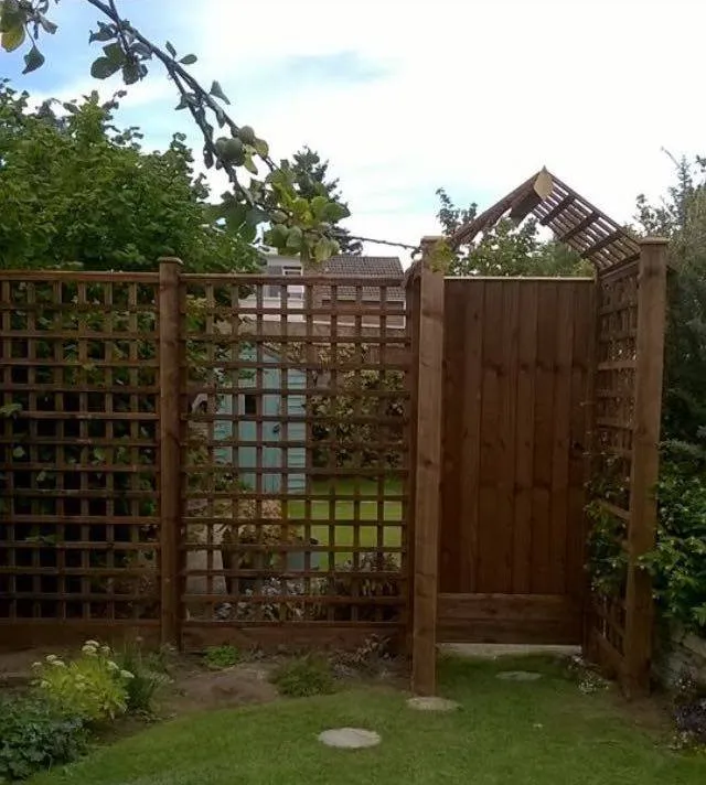 A wooden garden fence with a trellis design stands next to a gate. The fence encloses a garden area visible through the lattice. A lush garden and trees are visible in the background under a partly cloudy sky.