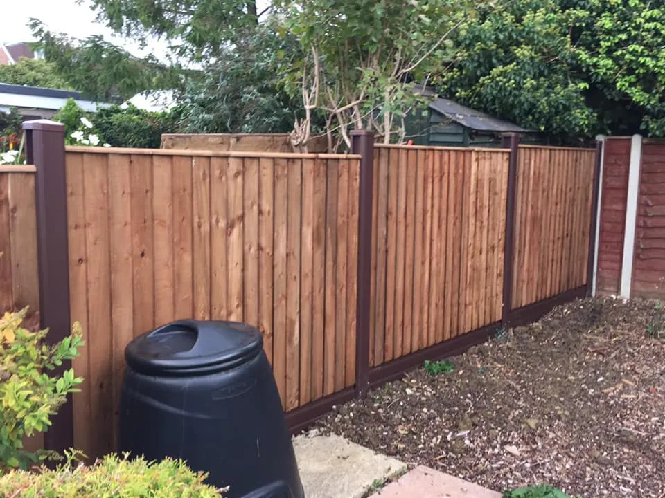 A wooden fence with brown posts runs along a garden, bordered by greenery and a small tree. In the foreground, a black compost bin rests on the ground, surrounded by soil and shrubs.