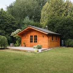 A small wooden cabin with a front porch sits amidst lush greenery. Surrounding the cabin are trees and bushes, and a well-kept lawn stretches out in front. Potted plants adorn the porch, adding to the rustic charm of the scene.