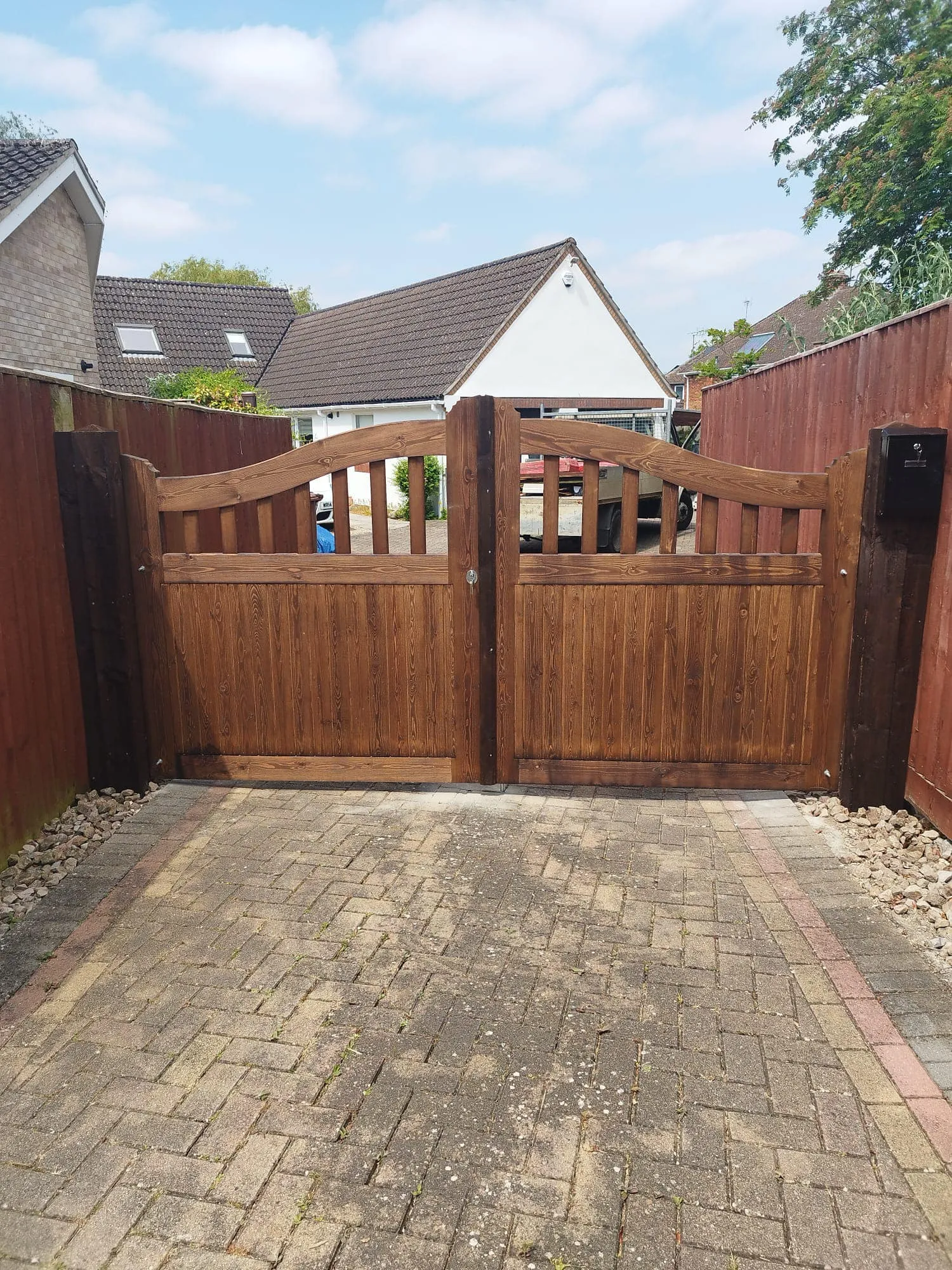A wooden gate with a curved design stands between two red fences in a residential driveway. The ground is paved with bricks, and there is a white house with a sloped roof in the background under a partly cloudy sky.