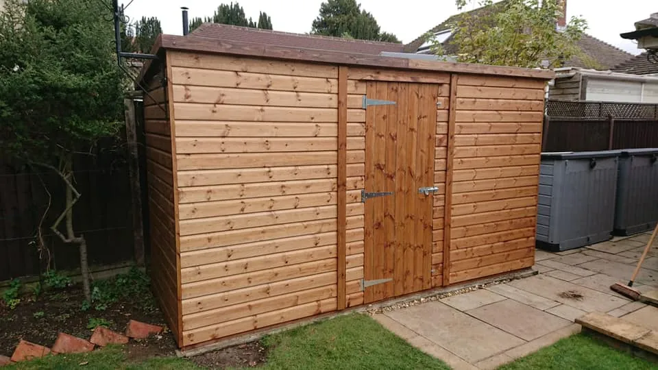 A wooden shed with a single door and metal hinges stands in a garden. It's bordered by a stone path and surrounded by grass, trees, and a wooden fence. Several gray plastic bins are visible in the background.