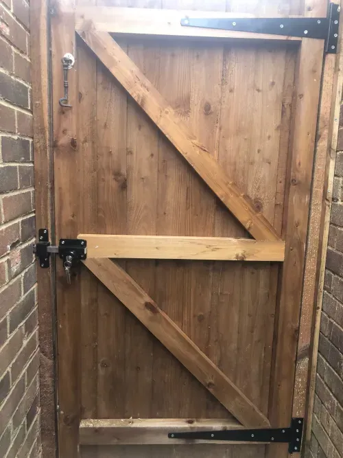 A sturdy wooden gate with a diagonal support beam, framed by brick walls on both sides. The gate features black metal hinges and a lock for security.